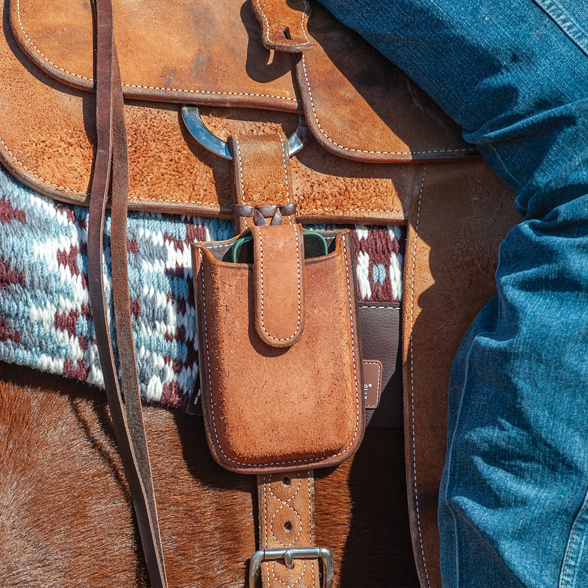 Natural roughout smartphone holder from Martin Saddlery hanging from a saddle beside a rider's leg. A phone sits inside the stitched pouch, which rests against a woven saddle blanket.