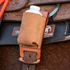 Natural roughout rope powder holder from Martin Saddlery on a saddle, seen up close with a white powder bottle inside. The holder hangs over the cinch strap below the tooled skirt.