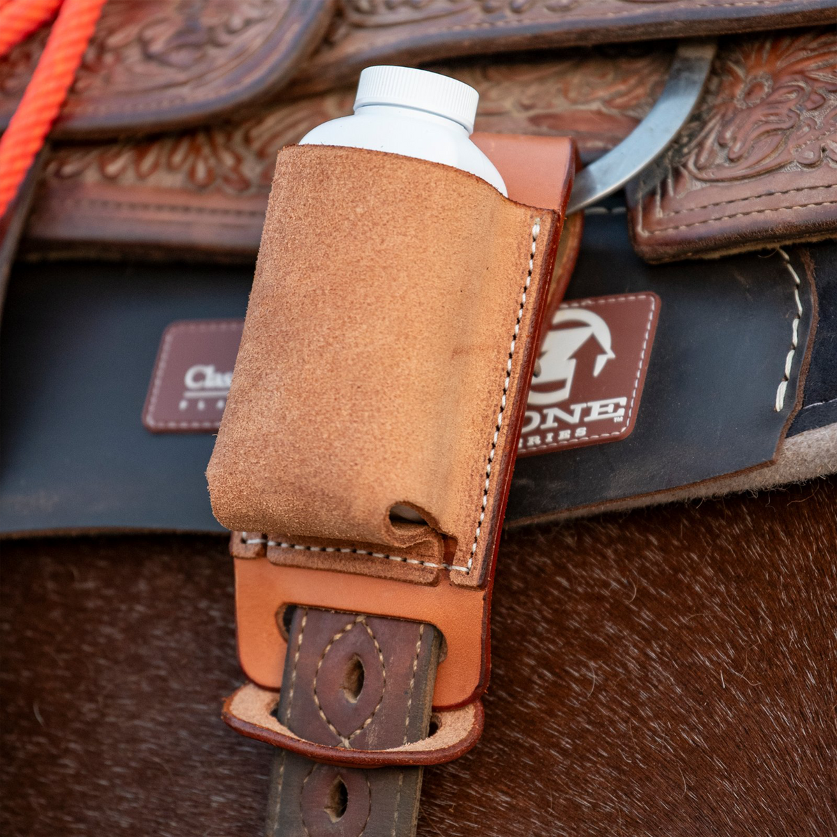 Natural roughout rope powder holder from Martin Saddlery on a saddle, seen up close with a white powder bottle inside. The holder hangs over the cinch strap below the tooled skirt.