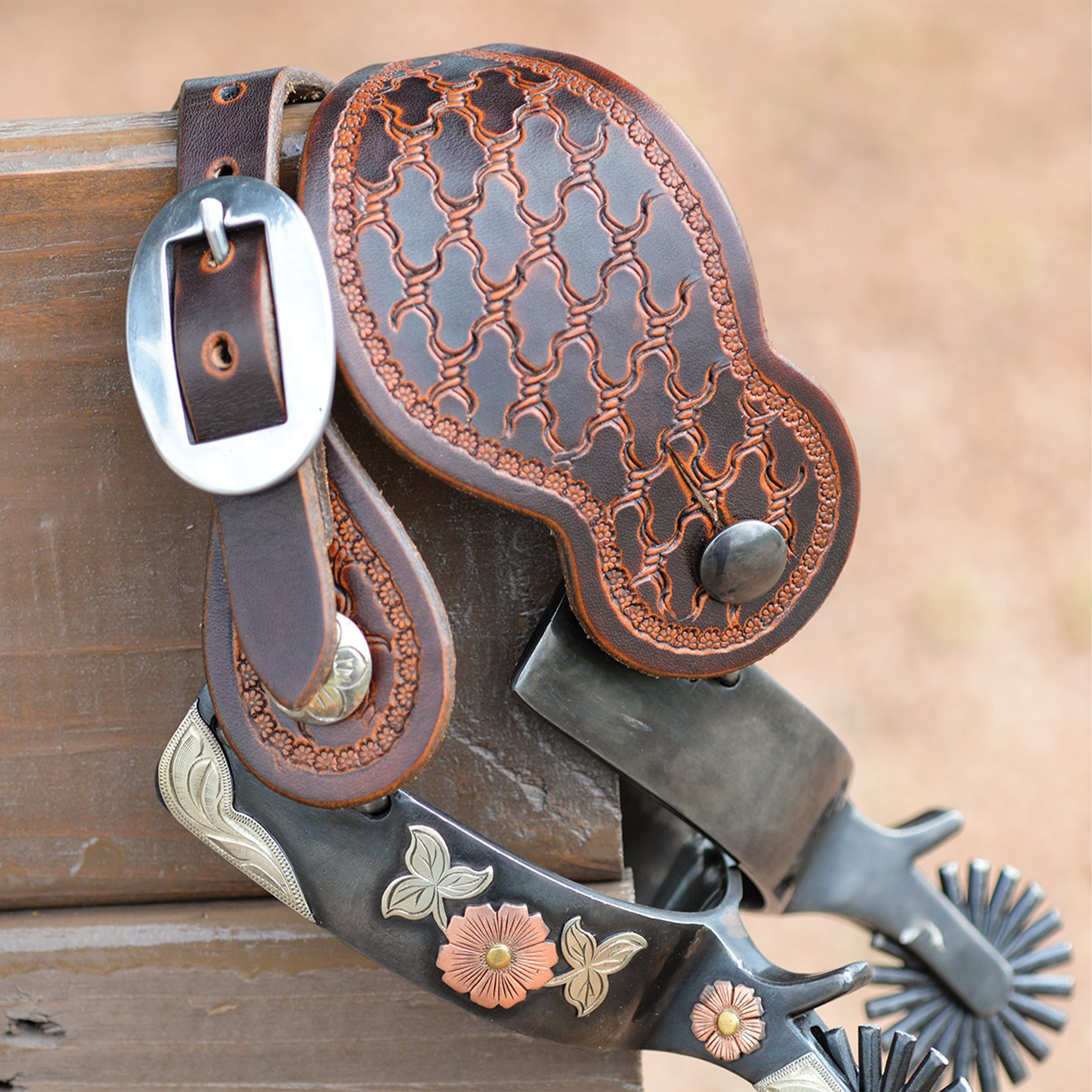 Weathered antique Tombstone spur strap with Twisted Wire tooling on a spur hooked over a wood rail. The tooled strap hangs beside its cart buckle above the engraved spur.
