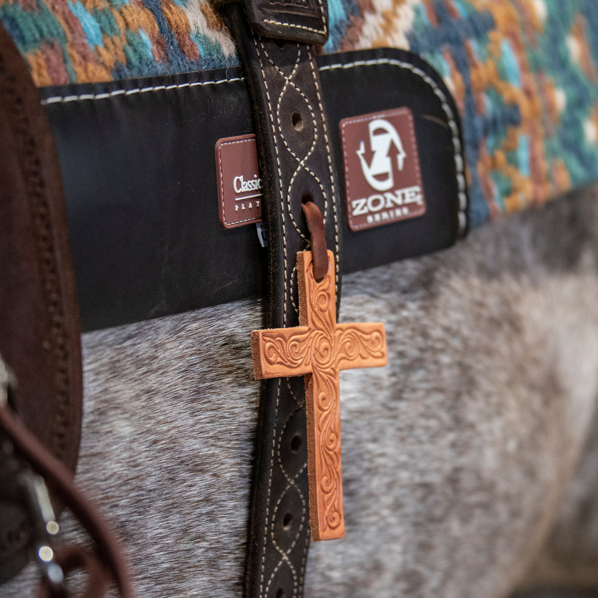 Natural tooled leather cross from Martin Saddlery hanging from a dark stitched strap on a saddled gray horse. The cross rests against the horse's side below a saddle pad and woven blanket.