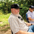 A man seated outdoors wearing the Original Hat x Martin Saddlery, a black trucker cap with a white shield logo patch on the front. The cap has a precurved bill and a mesh back.