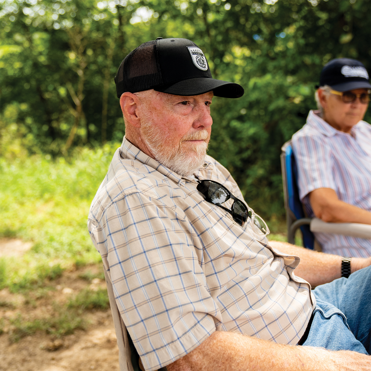 A man seated outdoors wearing the Original Hat x Martin Saddlery, a black trucker cap with a white shield logo patch on the front. The cap has a precurved bill and a mesh back.