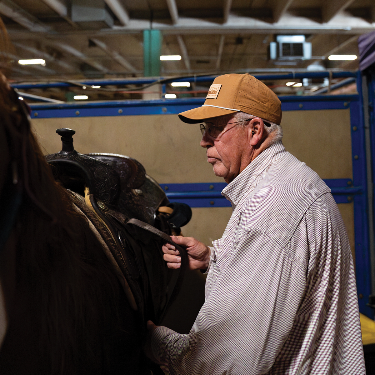 A man in a clay Performance Hat x Martin Saddlery stands beside a saddled horse in a barn. The clay snapback has a leather logo patch on the front.