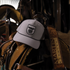 Silver Performance Hat x Martin Saddlery resting on a saddle in a dim tack room, with a tooled Martin Saddlery strap beside it. The cap has a black shield logo patch and a black rope across the bill.