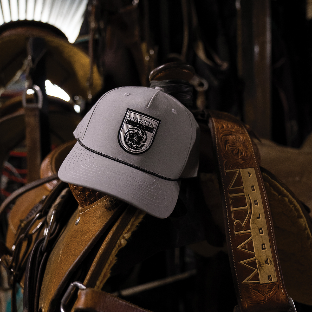Silver Performance Hat x Martin Saddlery resting on a saddle in a dim tack room, with a tooled Martin Saddlery strap beside it. The cap has a black shield logo patch and a black rope across the bill.