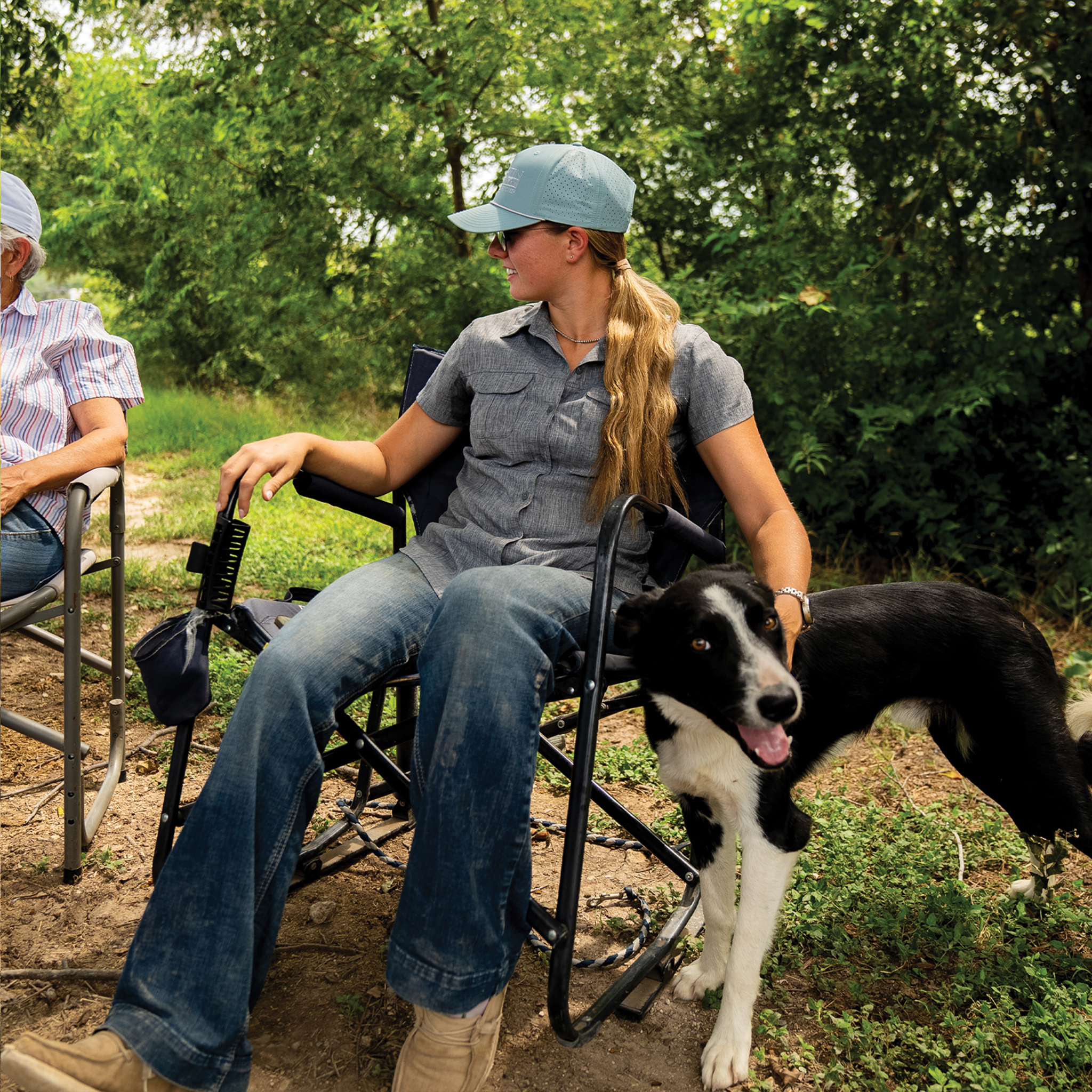 A woman in a smoke blue performance cap sits in a camp chair under trees, petting a black and white dog. She wears the Performance Hat x Martin Saddlery with a gray shirt and jeans.