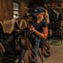 A woman in a navy Sport LT Hat x Martin Saddlery lifts a saddle in a tack room lined with saddle pads. The navy cap has a white embroidered logo on the front.