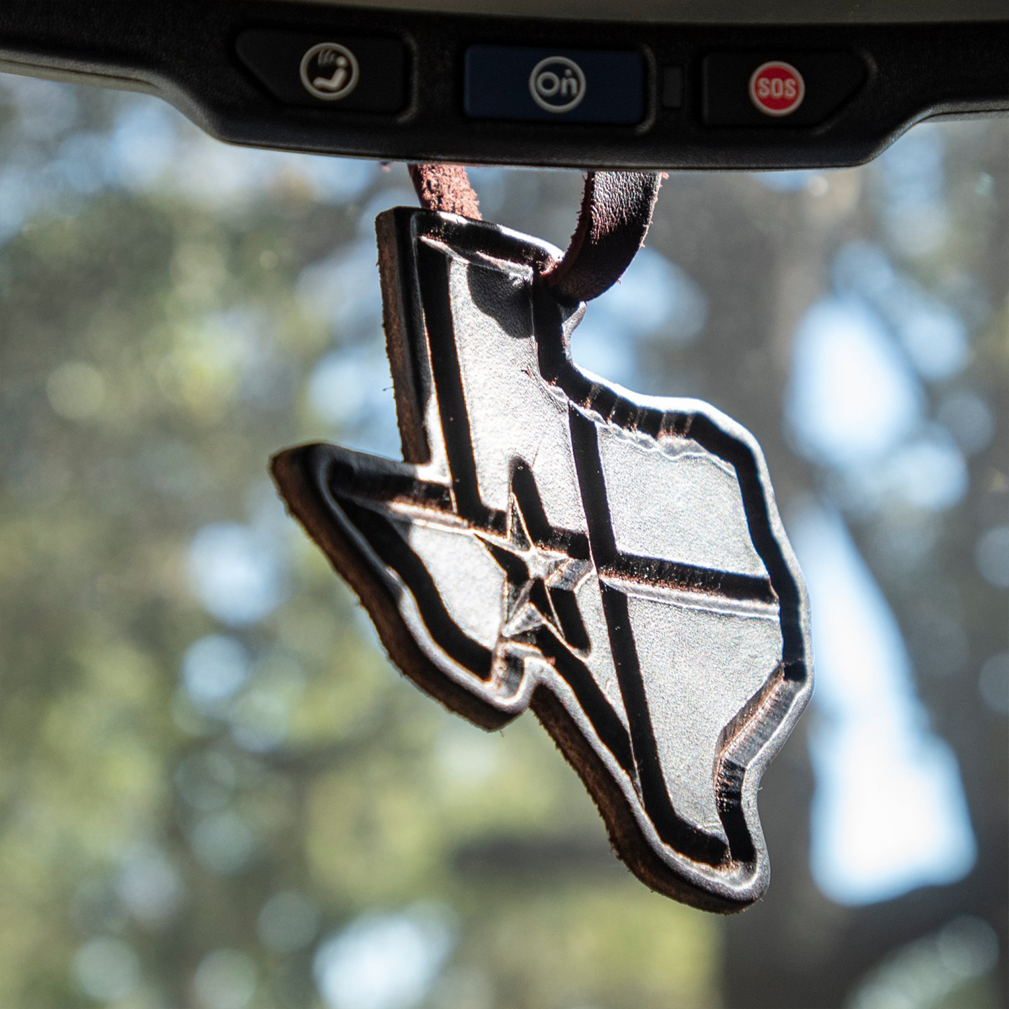 Texas shaped leather air freshener from Martin Saddlery hanging from a rearview mirror, with trees blurred behind. A star and flag lines are stamped into the dark leather.