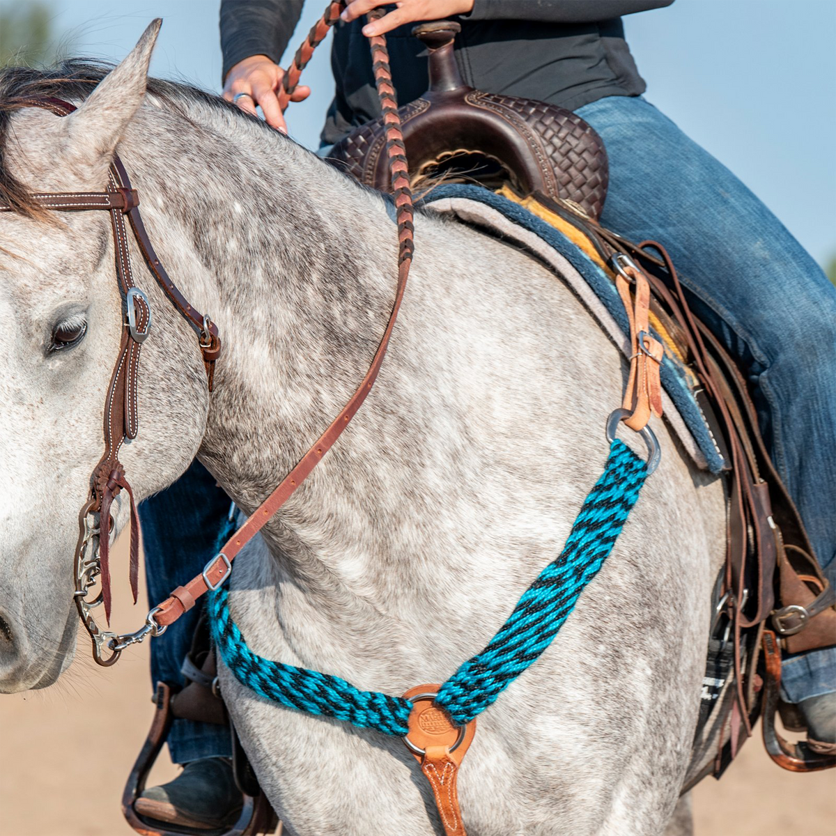 Martin Saddlery turquoise-black mohair breastcollar on a gray horse with a rider holding braided reins. The mohair strands cross the chest to a leather center piece with a tie-down strap, worn with a leather headstall.