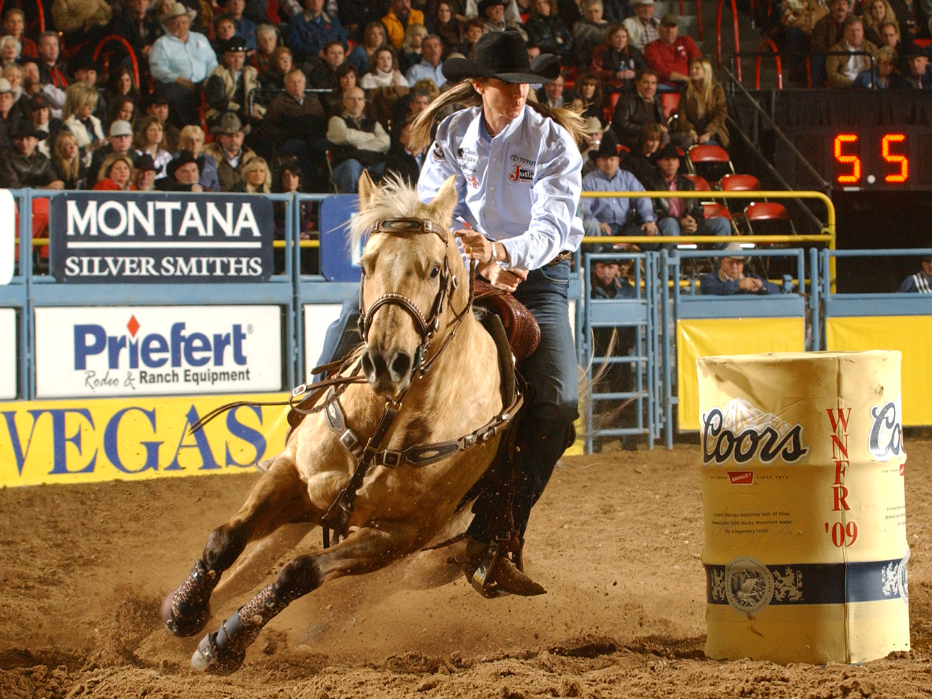 A cowgirl rides a palomino horse around a barrel in a rodeo arena, with a large crowd of spectators seated in the background and arena signage including Montana Silver Smiths, Priefert, and Vegas.