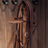 Natural floral tooled headstall hanging from a hook on a wooden tack room wall. The scroll tooled browband and cheek with chocolate-framed edges are visible beside a cart buckle.