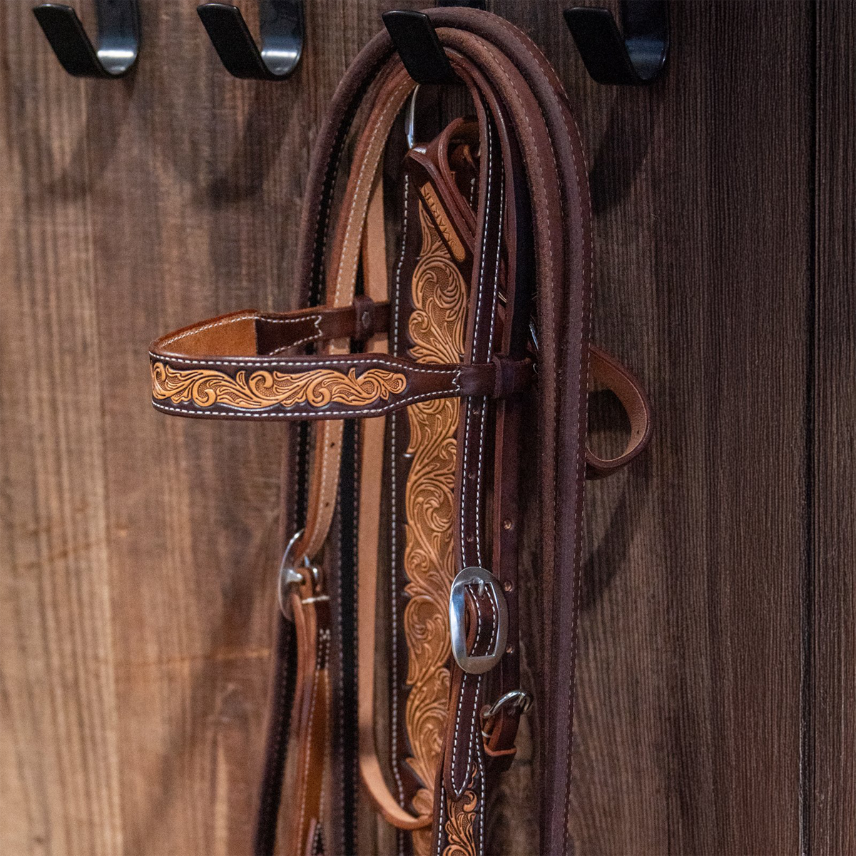 Natural floral tooled headstall hanging from a hook on a wooden tack room wall. The scroll tooled browband and cheek with chocolate-framed edges are visible beside a cart buckle.
