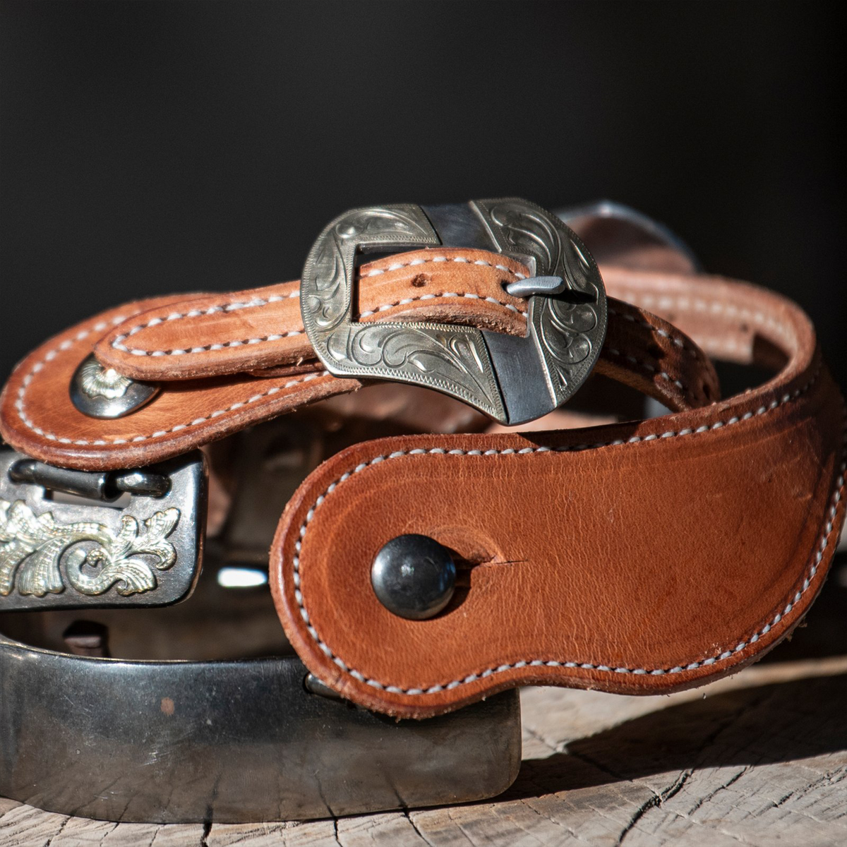 Natural harness spur strap on a spur resting on a wood block, seen up close against a dark background. White stitching edges the leather below the engraved Salinas buckle.