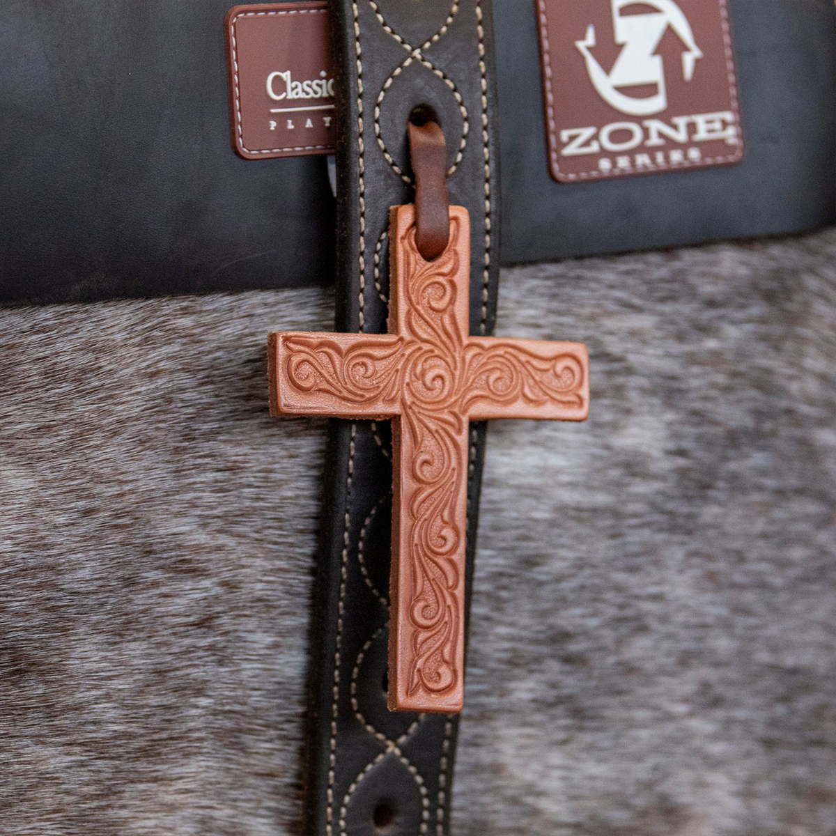 Natural leather cross saddle charm seen up close on a gray horse, tied to a dark strap with a leather string. Scroll tooling covers the face of the cross.