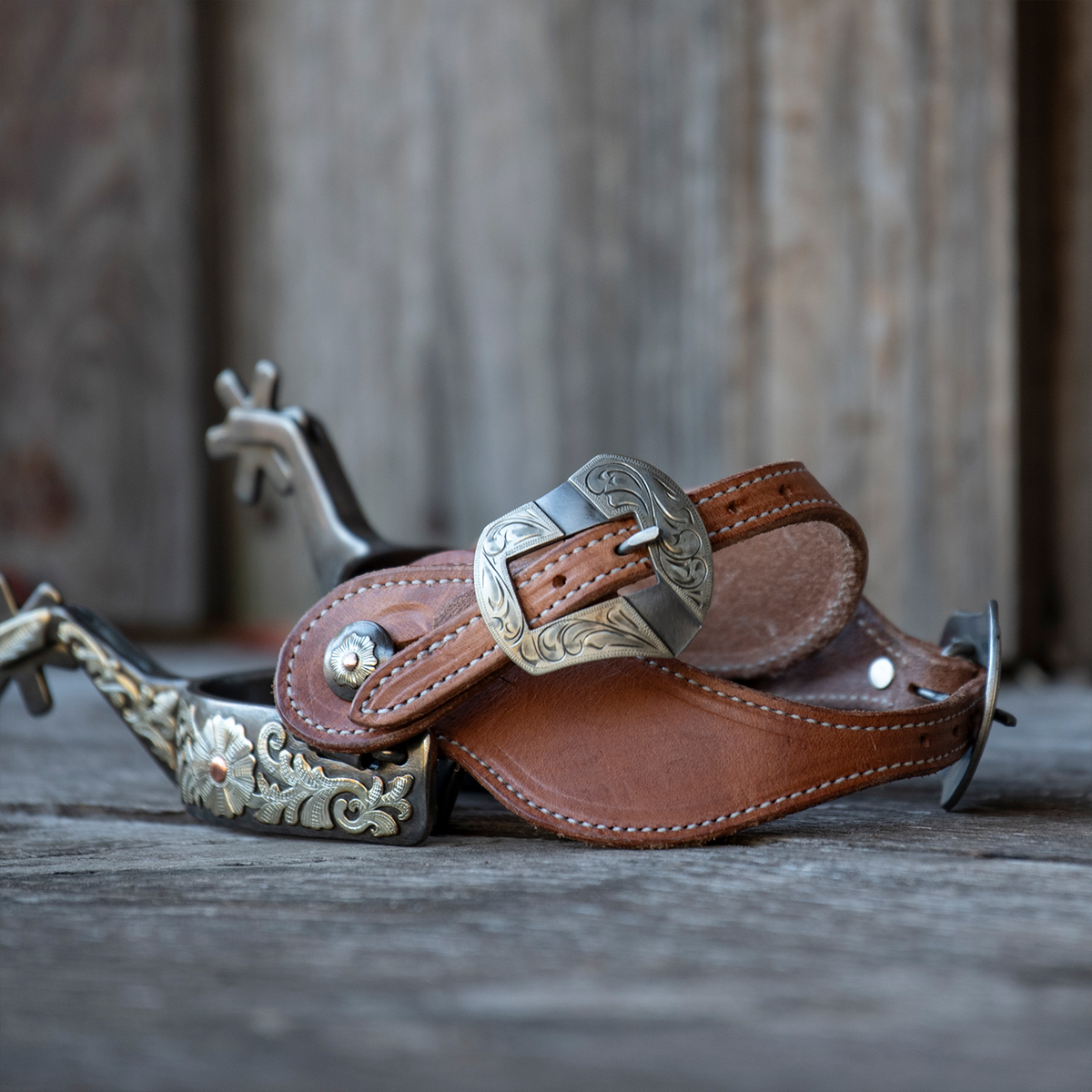 Spurs with natural harness spur straps sitting on weathered boards in front of a wood wall. The Salinas buckle sits on top of the stitched straps.