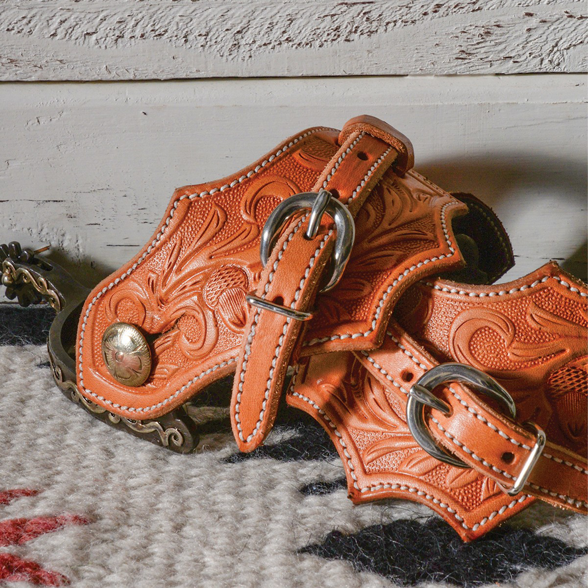Oak leaf spur straps on a pair of spurs resting on a woven blanket by a whitewashed wall. Acorn oak tooling covers the natural leather, with buckles across the top.
