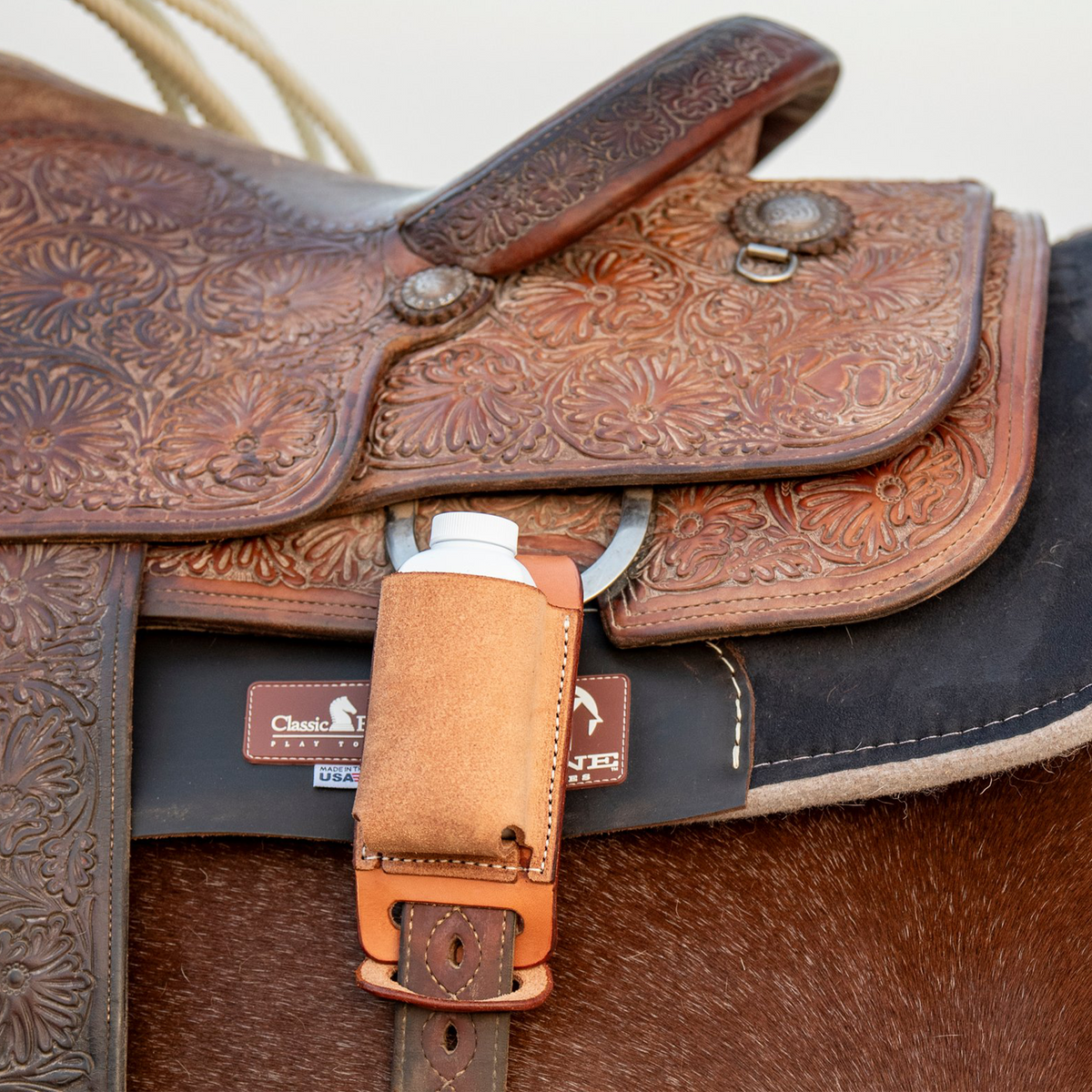 Tooled saddle on a sorrel horse with a natural roughout rope powder holder hanging from the back D-ring. A white powder bottle sits in the pocket above the cinch strap.