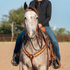 Gray horse facing the camera with a rider, wearing a natural scroll tooled breast collar. The tooled shoulder straps meet at a center ring on the chest above a tie-down strap, in an outdoor arena.