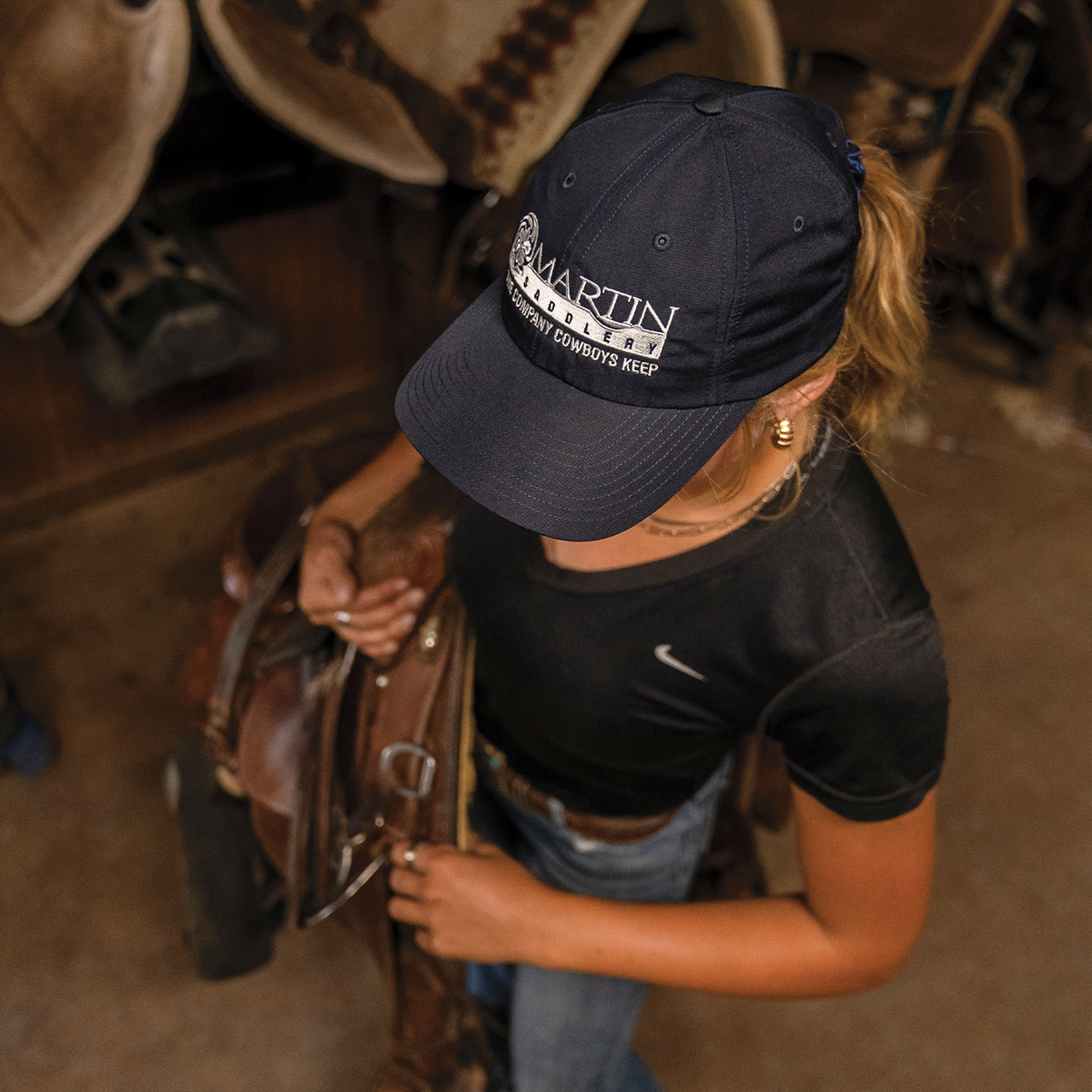 Navy sport cap with a white embroidered Martin Saddlery logo, seen from above on a woman holding a saddle. The relaxed-fit cap has a precurved bill.