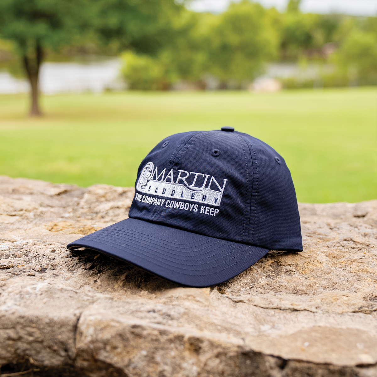 Navy six-panel hat resting on a stone wall with a lawn and trees behind it. The white embroidered Martin Saddlery logo reads The Company Cowboys Keep above the precurved bill.