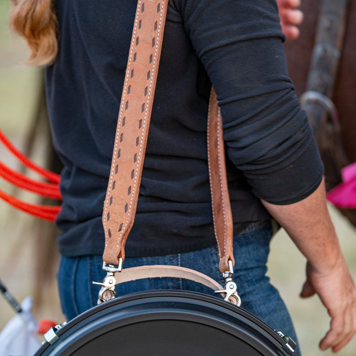Person carrying a rope can over one shoulder by a natural roughout buckstitched strap. Snaps join the strap to the can's handle, with a saddled horse behind.