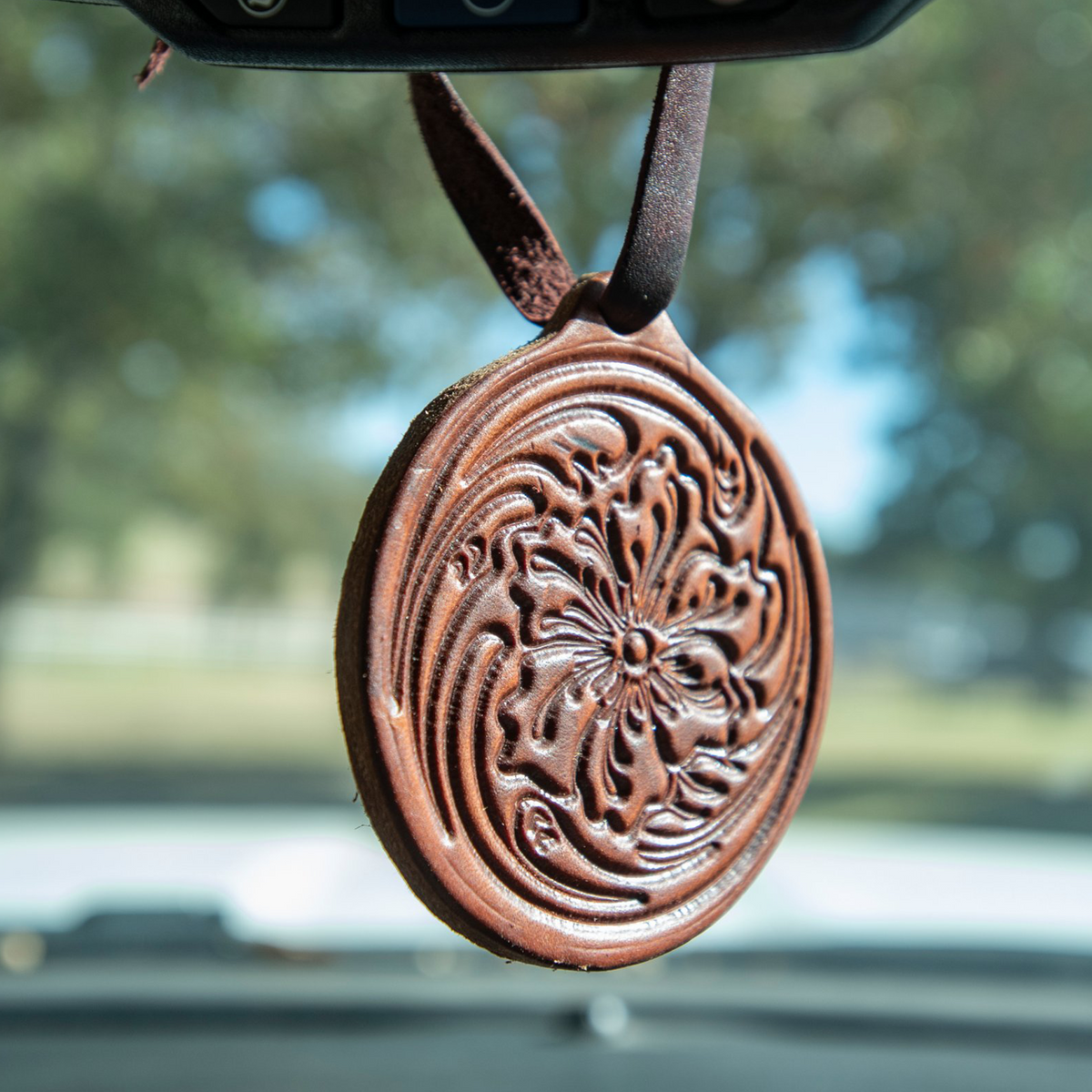 Brown leather air freshener with floral tooling hanging from a rearview mirror by a leather tie, above a dashboard. A tooled flower sits at the center of the round leather.