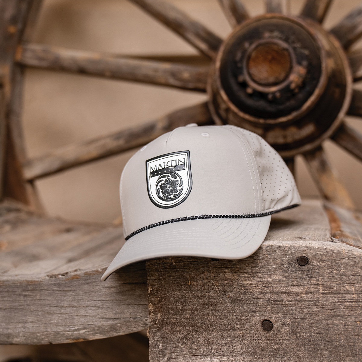 Silver rope hat with a black Martin Saddlery shield patch sitting on a weathered wooden bench in front of a wagon wheel. Laser-perforated side panels and a black rope across the bill are visible.