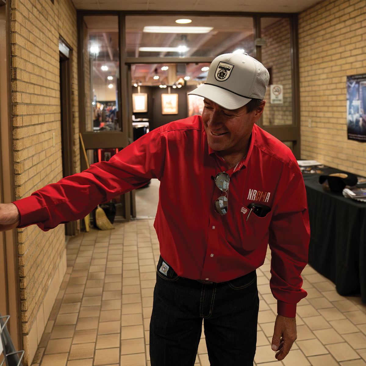 A man in a red shirt wearing a silver snapback cap with a black Martin Saddlery logo patch, standing in a hallway. The cap has a structured crown and precurved bill.