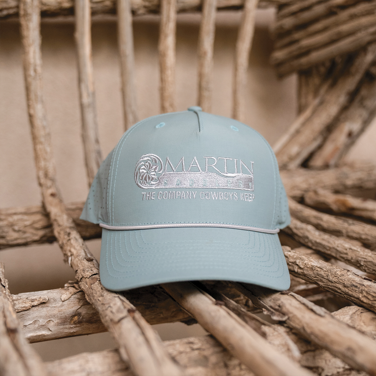 Smoke blue performance hat sitting on a chair made of bare wooden branches. The flat embroidered Martin Saddlery logo is centered on the front, with a light rope across the bill.