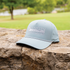 Smoke blue rope hat resting on a stone wall with a lawn and trees behind it. The Martin Saddlery logo and the words The Company Cowboys Keep are embroidered on the front above a rope across the bill.