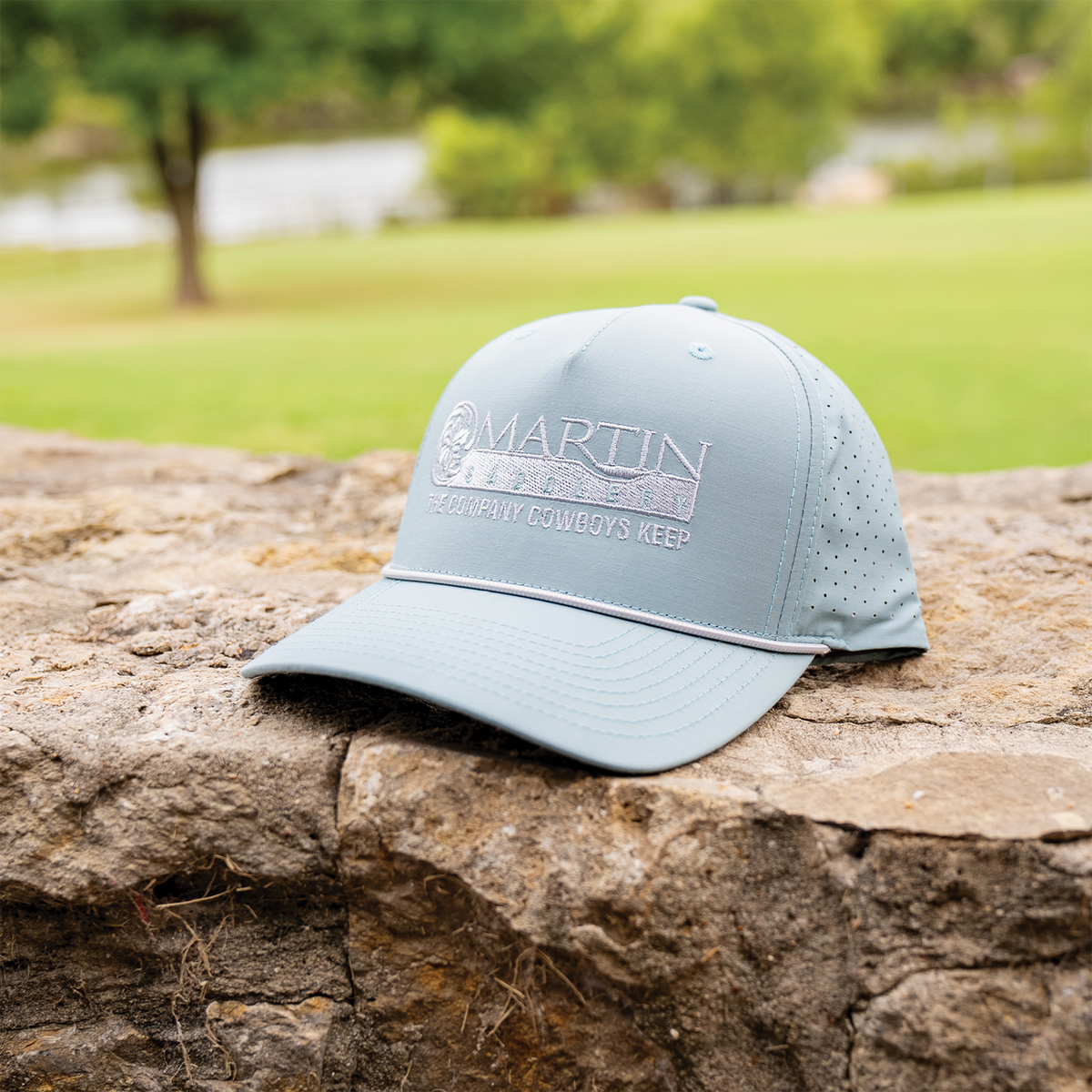 Smoke blue rope hat resting on a stone wall with a lawn and trees behind it. The Martin Saddlery logo and the words The Company Cowboys Keep are embroidered on the front above a rope across the bill.
