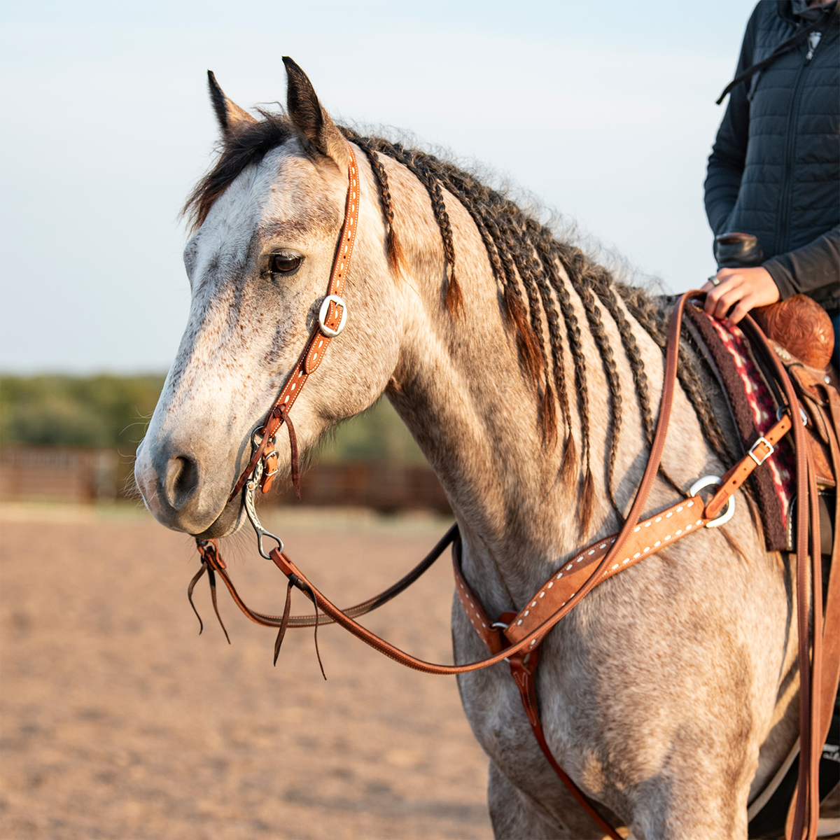 Gray horse with a braided mane and rider in an outdoor arena, wearing a western chestnut buckstitched headstall and a matching breastcollar. Latigo ties hold the bit below the cart buckle.