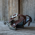 Pair of spurs with chocolate roughout spur straps sitting on weathered boards in front of a wood wall. The stitched straps loop over the spurs with the Prescott buckle on top.
