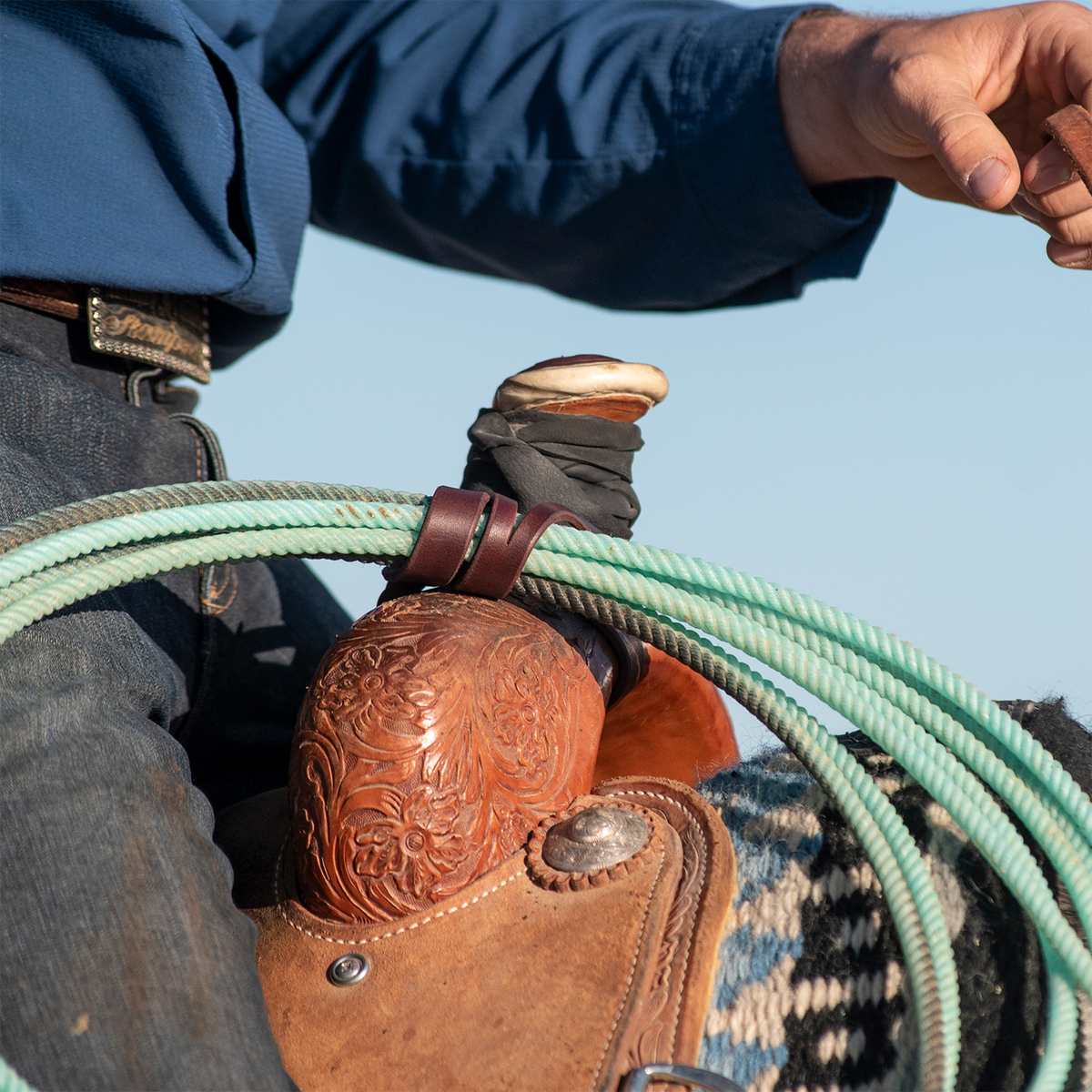 Rider in a blue shirt reaching forward beside a saddle horn where a latigo rope strap holds a green rope. The leather strap loops around the coils above the tooled swell.