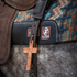 Tooled leather cross in natural leather hanging from a western saddle strap, with a roughout fender to one side. A woven saddle blanket and pad sit above the gray horse's side.