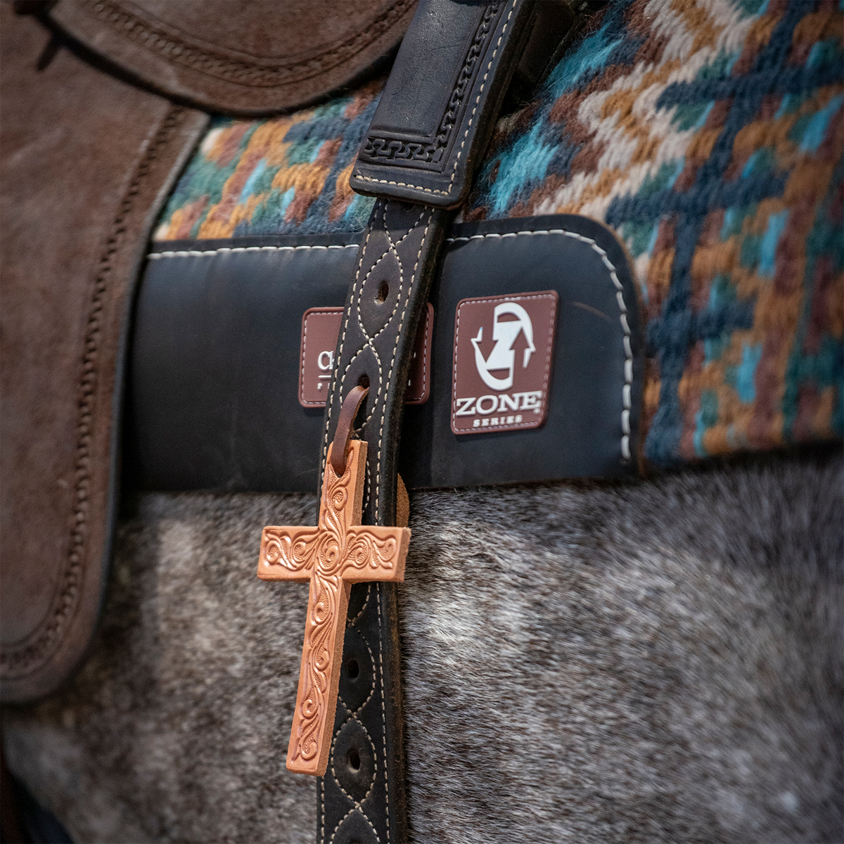 Tooled leather cross in natural leather hanging from a western saddle strap, with a roughout fender to one side. A woven saddle blanket and pad sit above the gray horse's side.