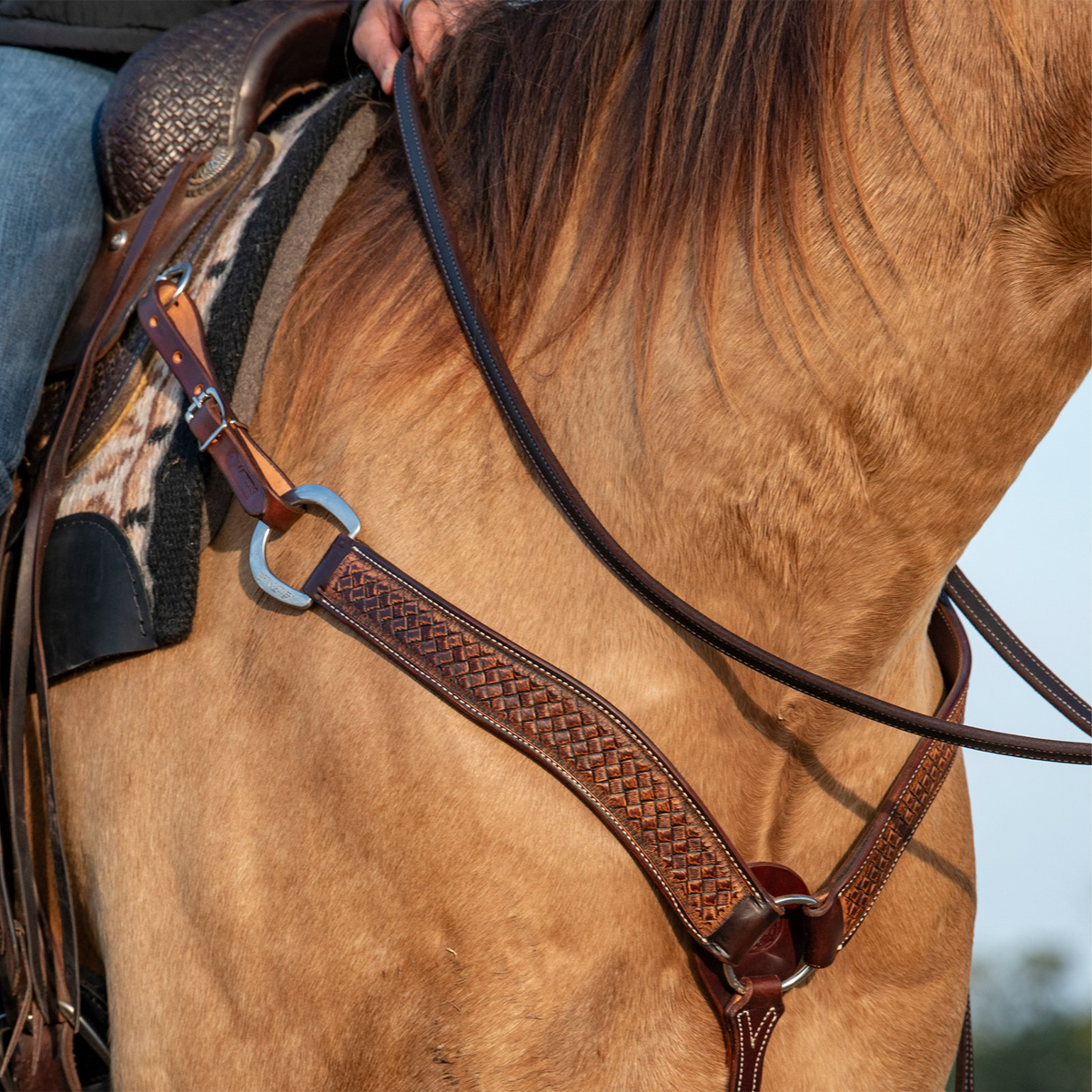 Western breast collar in weathered antique leather on a buckskin horse, seen at the shoulder. The weave stamped strap with stitched edges runs from a ring and buckle at the saddle down to the center of the chest.