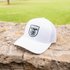 White trucker cap with a black Martin Saddlery shield logo resting on a stone wall, with a lawn behind it. The cap has a precurved bill and a white mesh back.
