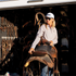 A woman in sunglasses and a white five panel hat carries a western saddle beside a trailer tack room. Bridles and tack hang in the shadows behind her.