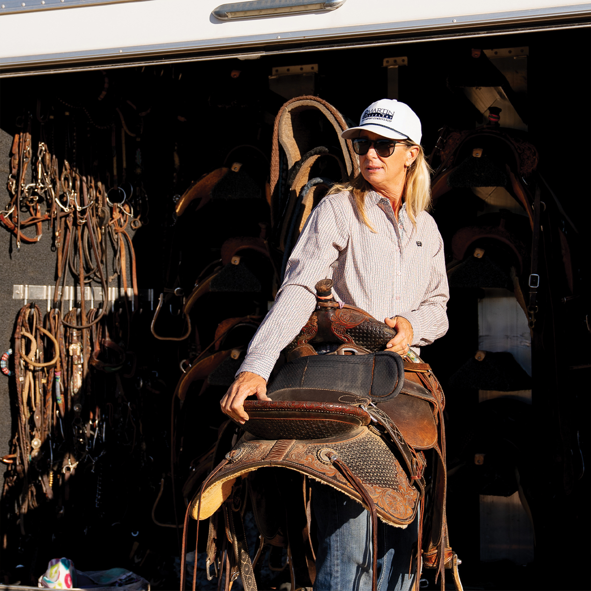 A woman in sunglasses and a white five panel hat carries a western saddle beside a trailer tack room. Bridles and tack hang in the shadows behind her.