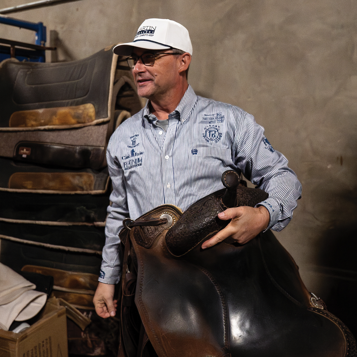 A man in a striped shirt and glasses wearing a white rope hat with a Martin Saddlery logo, holding a dark saddle. Saddle pads are stacked behind him.