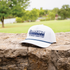 White rope hat resting on a stone wall with a lawn and trees behind it. A navy embroidered Martin Saddlery logo reads The Company Cowboys Keep above a braided navy rope on the bill.