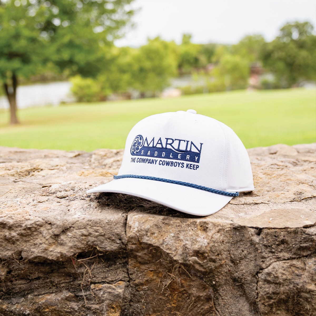 White rope hat resting on a stone wall with a lawn and trees behind it. A navy embroidered Martin Saddlery logo reads The Company Cowboys Keep above a braided navy rope on the bill.