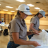 A woman in a white rope cap with a dark embroidered logo looks over papers at a table indoors. A second person in a light cap works beside her.