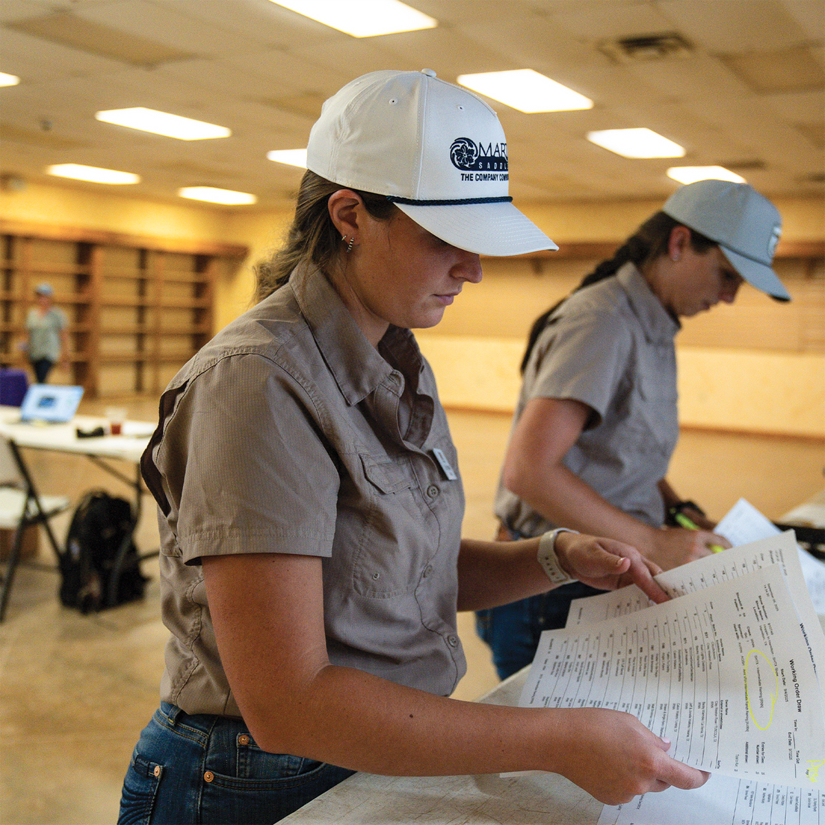 A woman in a white rope cap with a dark embroidered logo looks over papers at a table indoors. A second person in a light cap works beside her.