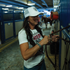 A woman in a white trucker cap adjusts a leather strap on a stall front in a barn. The cap has a black shield logo patch on the front and a white mesh back.