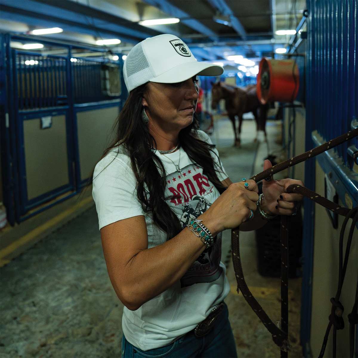 A woman in a white trucker cap adjusts a leather strap on a stall front in a barn. The cap has a black shield logo patch on the front and a white mesh back.